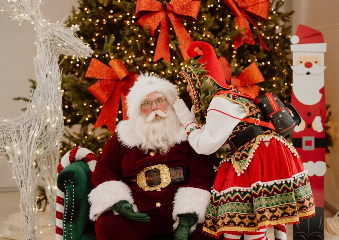 An elf whispering a secret to Santa in front of the tree