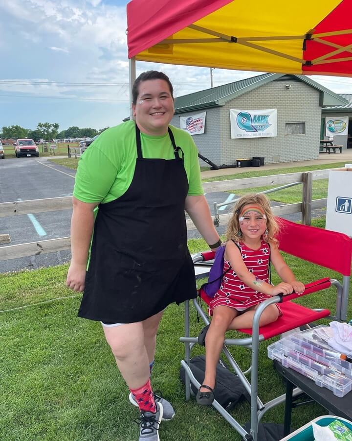Big Top Productions face painter posing with a smiling child showing off her rainbow design