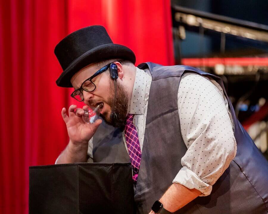 Adam in top hat performing a sleight-of-hand trick on stage