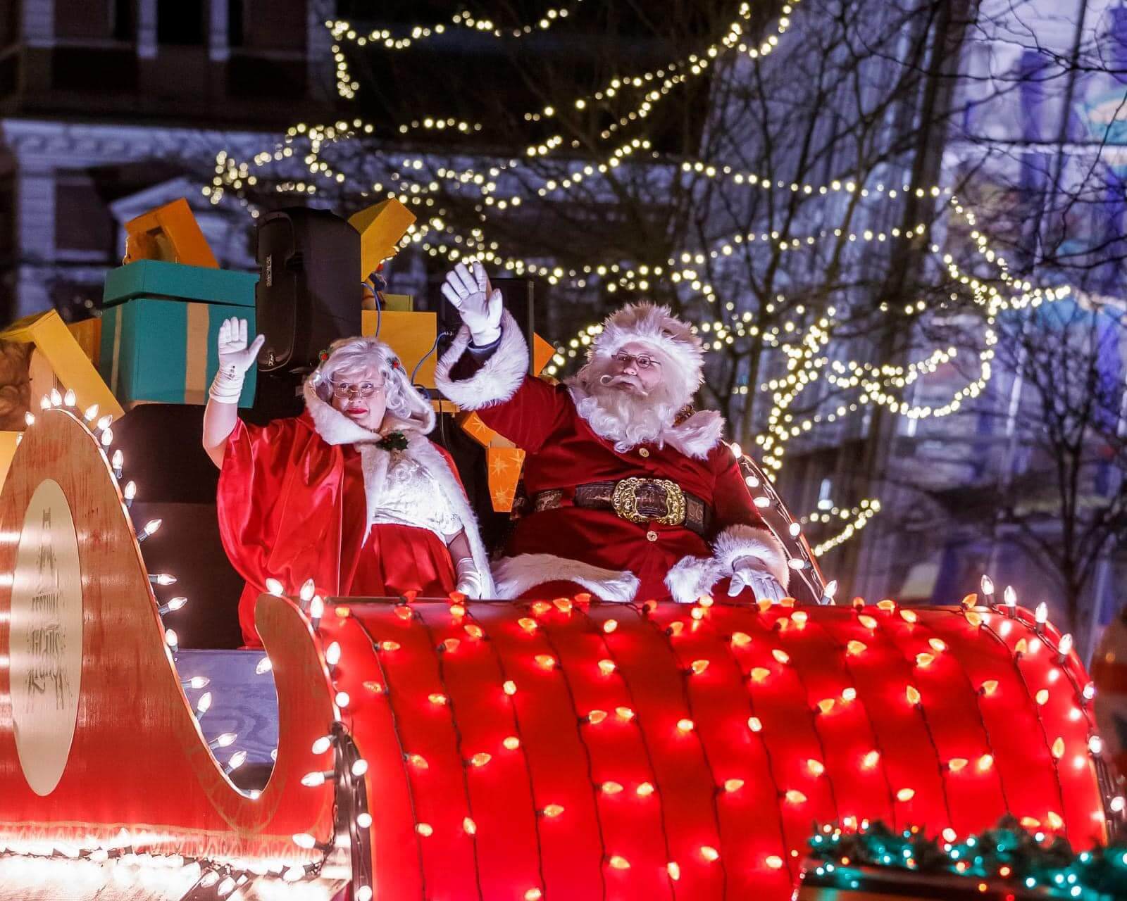 Santa and Mrs. Claus waving from a brightly lit sleigh float during a Christmas parade