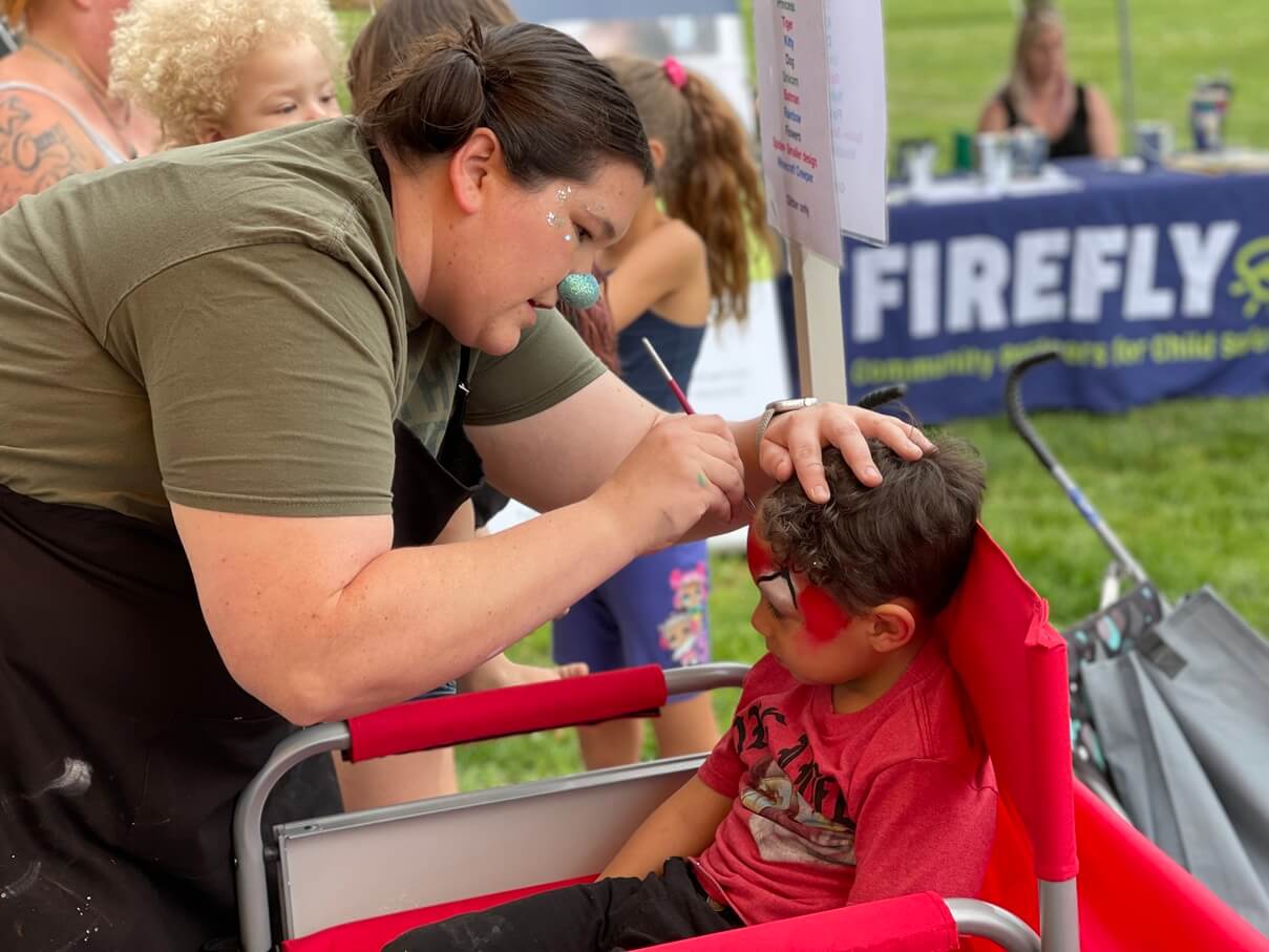 Face painter carefully applying a red design to a young boy's forehead